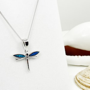 Puede incluir: Un collar de plata con un colgante de lib&eacute;lula con detalles de &oacute;palo azul. El collar est&aacute; sobre un fondo blanco con conchas marinas.