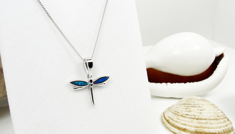 Puede incluir: Un collar de plata con un colgante de lib&eacute;lula con detalles de &oacute;palo azul. El collar est&aacute; sobre un fondo blanco con conchas marinas.