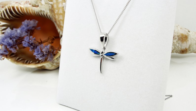 Puede incluir: Un collar con un colgante de lib&eacute;lula de plata con detalles de &oacute;palo azul. El collar est&aacute; sobre un fondo blanco.
