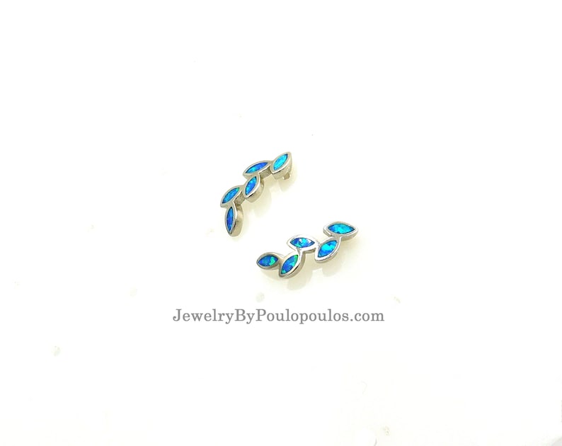 Op de afbeelding: Twee zilveren oormanchetten met een rij blauwe opaalstenen in de vorm van bladeren. De oormanchetten zijn gebogen en hebben een delicaat ontwerp. JewelryByPoulopoulos.com