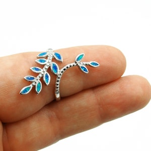 May include: A silver-toned ring featuring a branch design with blue opal leaf accents. The ring is held up by a finger against a white background. The leaves have a blue and teal color.