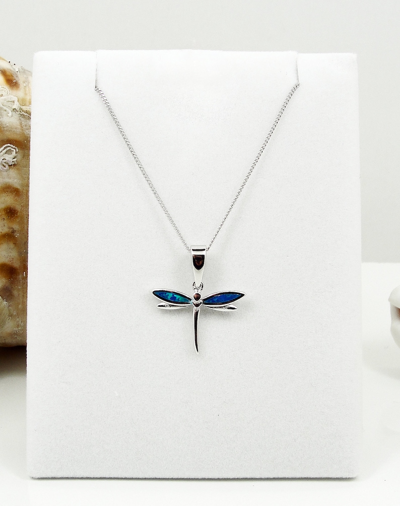 Puede incluir: Un collar de plata con un colgante de lib&eacute;lula con alas de &oacute;palo azul y un acento de piedra roja. El collar est&aacute; en un expositor blanco.