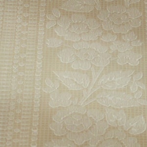 Puede incluir: Primer plano de una tela beige con un patr&oacute;n floral blanco. La tela tiene una superficie texturizada y un brillo sutil.