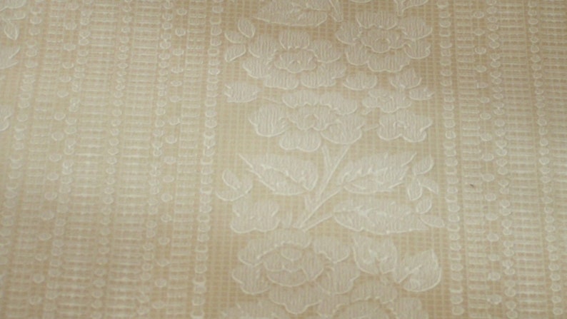 Puede incluir: Primer plano de una tela beige con un patr&oacute;n floral blanco. La tela tiene una superficie texturizada y un brillo sutil.