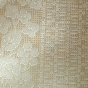 Puede incluir: Una tela beige con un patr&oacute;n en relieve blanco de flores y hojas. La tela tiene un sutil patr&oacute;n de cuadr&iacute;cula.