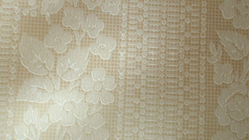 Puede incluir: Una tela beige con un patr&oacute;n en relieve blanco de flores y hojas. La tela tiene un sutil patr&oacute;n de cuadr&iacute;cula.