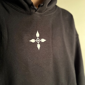 May include: Black hoodie with a white emblem on the chest. The hoodie has a drawstring hood and a simple, minimalist design. The emblem is a stylized cross or compass-like shape.