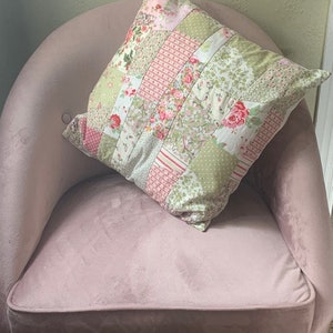 May include: A pink velvet armchair with a patchwork floral pillow. The pillow has a mix of pink, green, and white floral patterns.