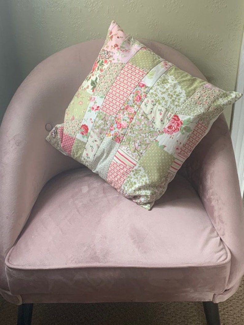May include: A pink velvet armchair with a patchwork floral pillow. The pillow has a mix of pink, green, and white floral patterns.