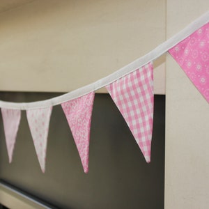 May include: A pink and white fabric bunting banner with various patterns, including gingham and floral designs. The pennants are strung on a white ribbon, creating a decorative accent. The banner is suitable for parties and celebrations.