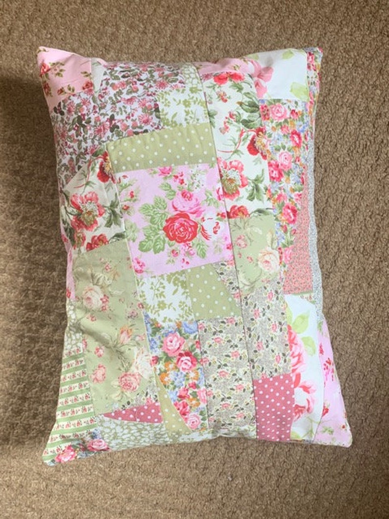 May include: A patchwork pillow with a floral design in shades of pink, green, and white. The pillow is made of different fabrics and has a quilted texture.