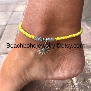 May include: Yellow and green beaded ankle bracelet with a silver sunflower charm. The bracelet has two silver beads with a swirl design.