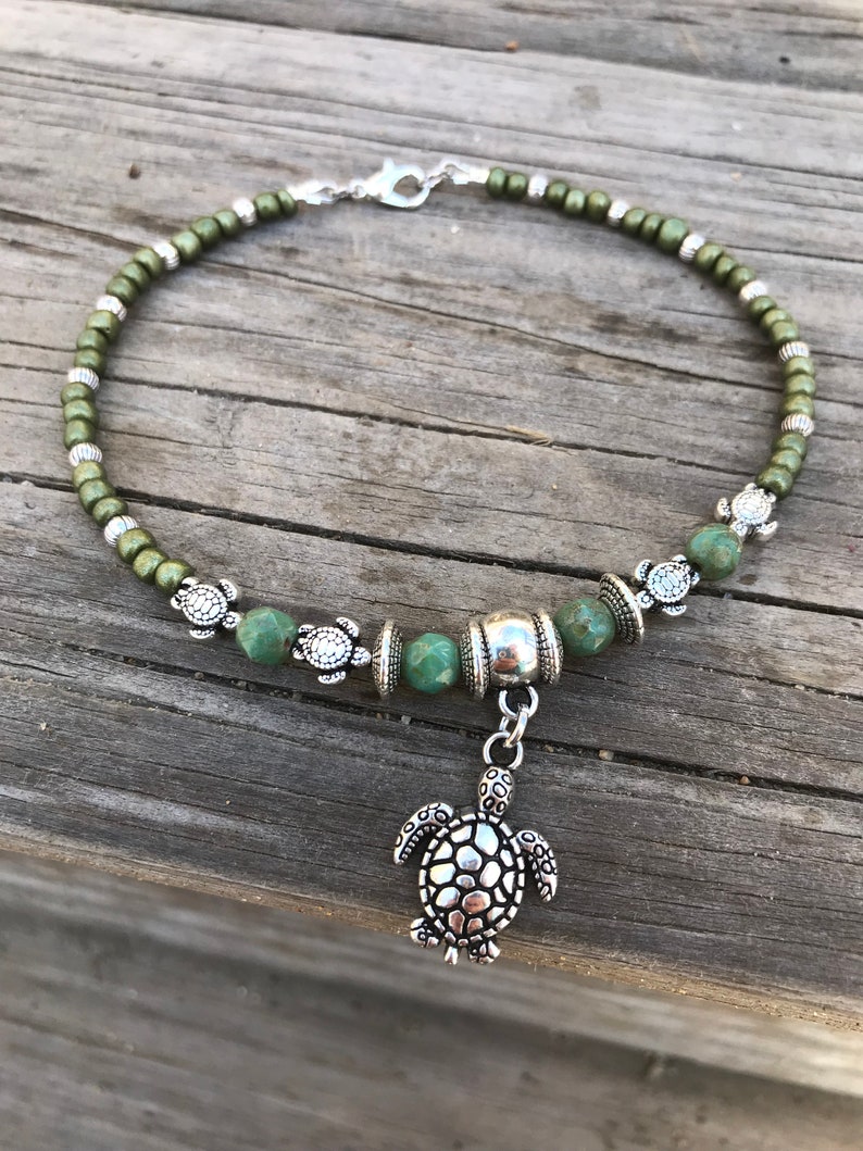 May include: Green beaded ankle bracelet with a silver turtle charm. The bracelet has small silver turtle beads and a silver bead with a textured surface.