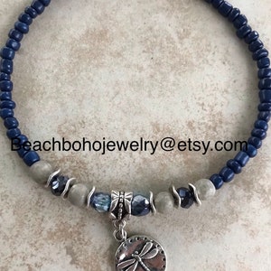 May include: A blue and gray beaded bracelet with a silver dragonfly charm. The bracelet has a silver clasp and is made with small, round beads.