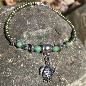 May include: A green and silver beaded necklace with a silver turtle charm. The necklace is made with small, round beads and has a silver toggle clasp.