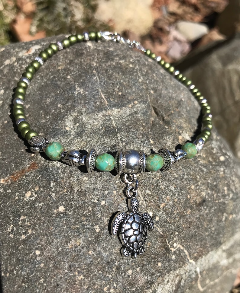 May include: A green and silver beaded necklace with a silver turtle charm. The necklace is made with small, round beads and has a silver toggle clasp.