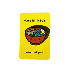 Puede incluir: Un pin de esmalte amarillo con el texto "mochi kids" y "enamel pin". El pin presenta una ilustración de dibujos animados de un cuenco rojo de ramen con una cara sonriente.