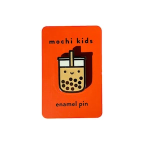May include: A gold enamel pin featuring a cartoon illustration of a boba tea cup with a smiling face and a straw. The pin is on a red card with the text "mochi kids" and "enamel pin".