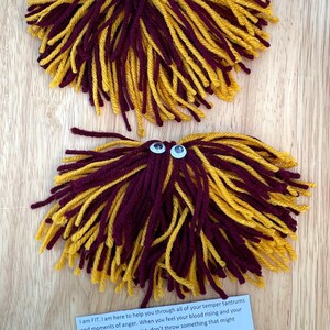 May include: Two fuzzy yarn stress balls with googly eyes. The yarn is maroon and gold. The stress balls have a white tag attached with the text "THROW A FIT" and instructions to use the stress ball when feeling angry.
