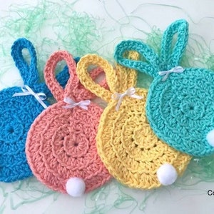 May include: Four crocheted bunny-shaped ornaments in blue, peach, yellow, and teal colors. Each ornament has a white pom-pom tail and a white ribbon bow. The ornaments are arranged on a bed of green shredded paper. Color Combo #2