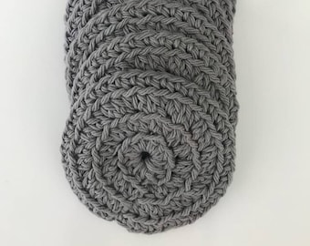 May include: A stack of gray, crocheted coasters. Each coaster is round with a spiral design in the center. The coasters are made of a thick, textured yarn and are a solid gray color. The coasters are stacked on top of each other.