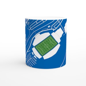 May include: White ceramic mug featuring a detailed blue and white illustration of a soccer stadium. The stadium's green field and surrounding structures are depicted with precision, set against a vibrant blue background.