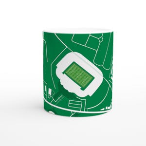 Puede incluir: Taza de cerámica blanca con una ilustración verde y blanca de un estadio. El diseño muestra una vista aérea detallada del estadio, incluyendo el campo y los edificios circundantes. La taza tiene un acabado brillante y un asa estándar.