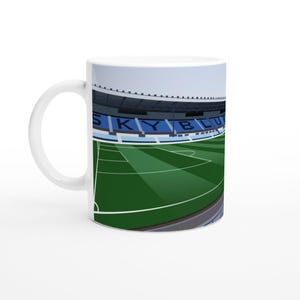 Può includere: Tazza in ceramica bianca con un design a stadio. La tazza presenta un campo verde, posti a sedere blu e la scritta "SKY BLUE". La tazza ha un manico curvo. Il design è un'illustrazione stilizzata.