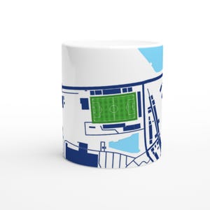 May include: White ceramic mug featuring a detailed map design of a sports stadium. The design includes a green field, blue accents, and dark blue outlines. The mug is cylindrical and sits on a white surface.