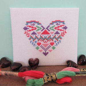 Puede incluir: Un proyecto de punto de cruz blanco con un dise&ntilde;o de coraz&oacute;n colorido con patrones geom&eacute;tricos en rojo, azul, morado y rosa. El coraz&oacute;n est&aacute; centrado en la tela. Varias piedras de vidrio en forma de coraz&oacute;n y madejas de hilo de bordar de varios colores est&aacute;n en primer plano.