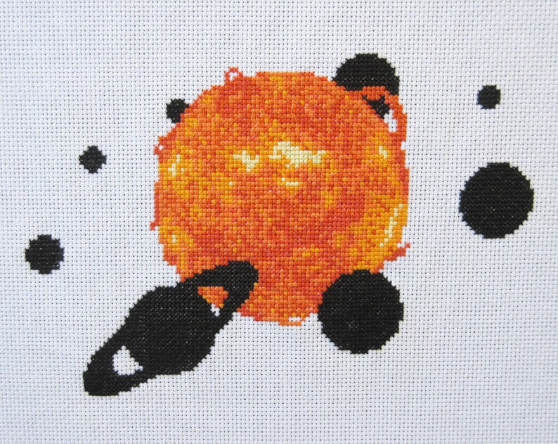May include: A cross-stitch design of a large orange sun with smaller black circles around it, representing planets.
