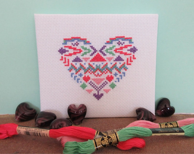 Puede incluir: Un proyecto de punto de cruz blanco con un dise&ntilde;o de coraz&oacute;n colorido con patrones geom&eacute;tricos en rojo, azul, morado y rosa. El coraz&oacute;n est&aacute; centrado en la tela. Varias piedras de vidrio en forma de coraz&oacute;n y madejas de hilo de bordar de varios colores est&aacute;n en primer plano.