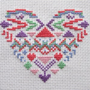 Puede incluir: Un dise&ntilde;o de coraz&oacute;n en punto de cruz en varios colores, incluyendo rojo, morado, verde y azul, sobre un fondo de tela blanca. El coraz&oacute;n presenta patrones geom&eacute;tricos y elementos florales, creando una est&eacute;tica de arte popular.