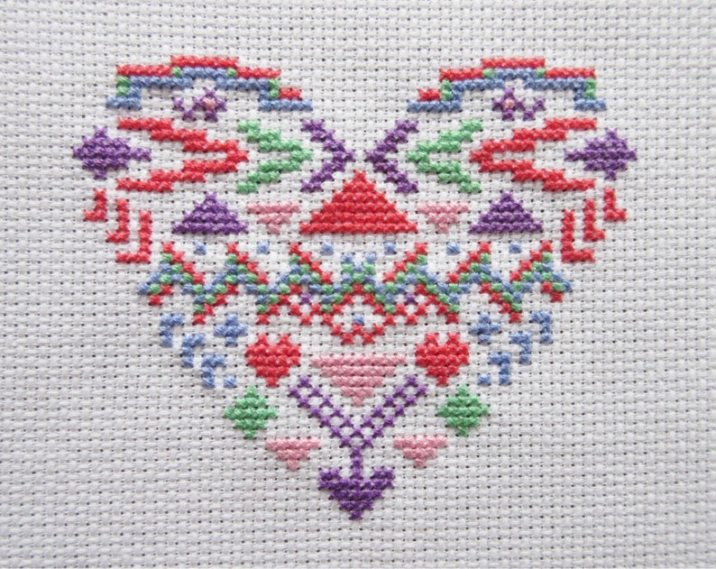 Puede incluir: Un dise&ntilde;o de coraz&oacute;n en punto de cruz en varios colores, incluyendo rojo, morado, verde y azul, sobre un fondo de tela blanca. El coraz&oacute;n presenta patrones geom&eacute;tricos y elementos florales, creando una est&eacute;tica de arte popular.