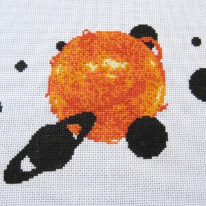 May include: Cross-stitch artwork depicting a red and orange sun with a black planet and rings orbiting around it. The background is white with black dots.