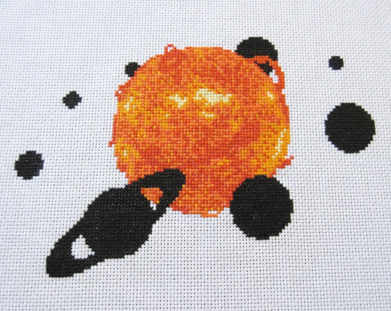 May include: Cross-stitch artwork depicting a red and orange sun with a black planet and rings orbiting around it. The background is white with black dots.