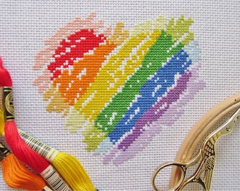 Puede incluir: Un patrón de punto de cruz en forma de corazón arcoíris sobre tela Aida blanca. El corazón está hecho de puntos de diferentes colores, incluyendo rosa, rojo, naranja, amarillo, verde, azul, morado y blanco.