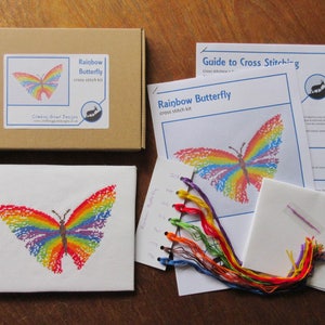 May include: A cross stitch kit featuring a rainbow butterfly design. The kit includes a piece of Aida cloth, embroidery floss in various colors, a needle, and instructions. The kit is packaged in a brown box with a label that says "Rainbow Butterfly cross stitch kit".