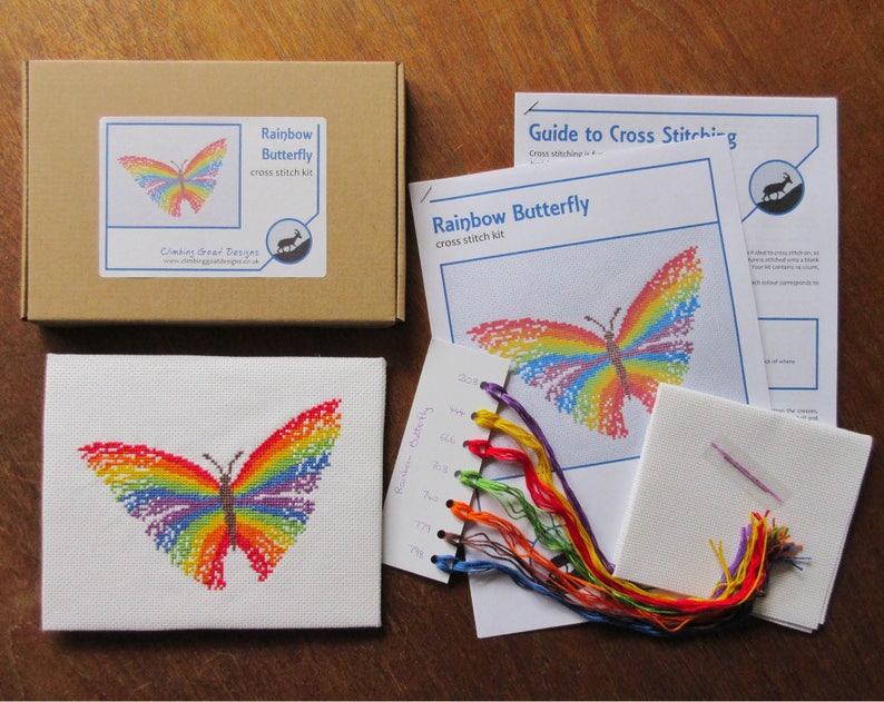 May include: A cross stitch kit featuring a rainbow butterfly design. The kit includes a piece of Aida cloth, embroidery floss in various colors, a needle, and instructions. The kit is packaged in a brown box with a label that says "Rainbow Butterfly cross stitch kit".