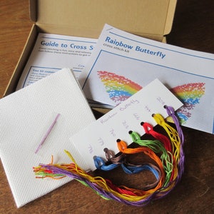 May include: A cross stitch kit featuring a rainbow butterfly design. The kit includes a piece of Aida cloth, embroidery floss in various colors, a needle, and instructions. The floss colors are labeled with DMC numbers: 798, 779, 740, 703, 666, 434, and 208.