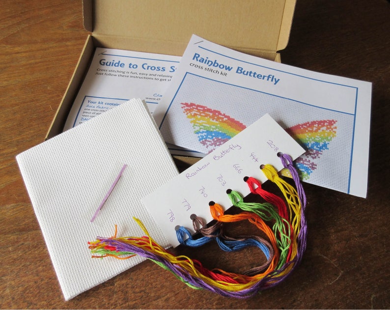 May include: A cross stitch kit featuring a rainbow butterfly design. The kit includes a piece of Aida cloth, embroidery floss in various colors, a needle, and instructions. The floss colors are labeled with DMC numbers: 798, 779, 740, 703, 666, 434, and 208.