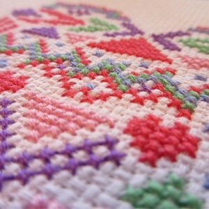 Puede incluir: Primer plano de un bordado de punto de cruz con un dise&ntilde;o floral. El bordado presenta una variedad de colores, incluyendo rojo, rosa, morado, verde y azul, sobre un fondo de tela blanca. Las puntadas crean un patr&oacute;n texturizado y detallado.