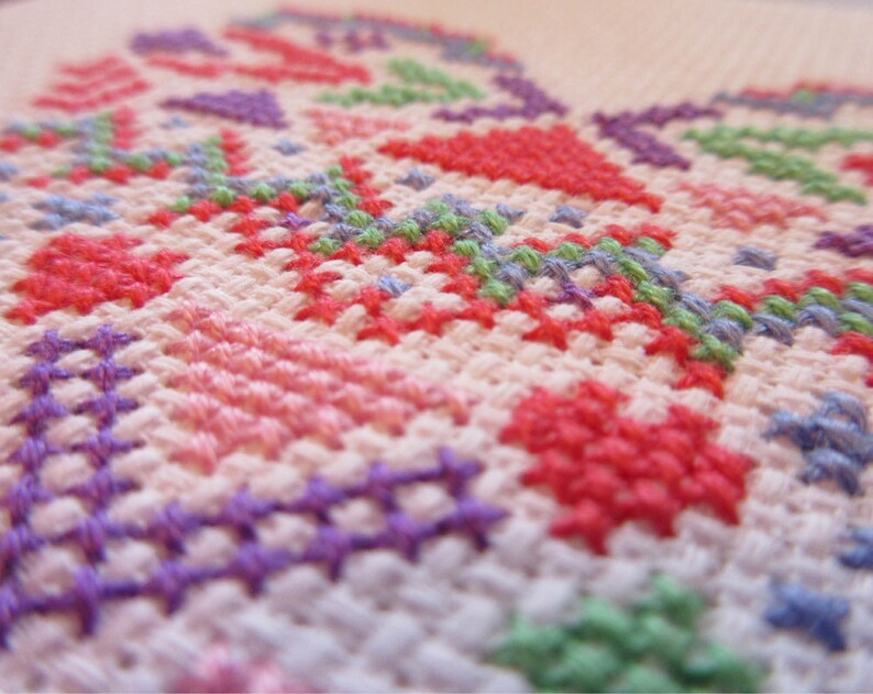 Puede incluir: Primer plano de un bordado de punto de cruz con un dise&ntilde;o floral. El bordado presenta una variedad de colores, incluyendo rojo, rosa, morado, verde y azul, sobre un fondo de tela blanca. Las puntadas crean un patr&oacute;n texturizado y detallado.