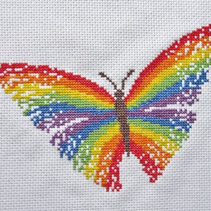 May include: A rainbow-colored butterfly cross-stitch pattern on white fabric. The butterfly is stitched with red, orange, yellow, green, blue, indigo, and violet thread.