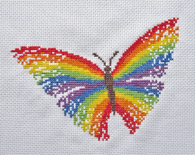 May include: A rainbow-colored butterfly cross-stitch pattern on white fabric. The butterfly is stitched with red, orange, yellow, green, blue, indigo, and violet thread.