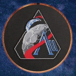 Puede incluir: Arte de bordado a punto de cruz con un diseño geométrico que presenta un triángulo y una letra "A" estilizada. El diseño incorpora elementos rojos, azules y grises sobre un fondo negro, enmarcado en un aro de madera. El fondo es un cielo estrellado.