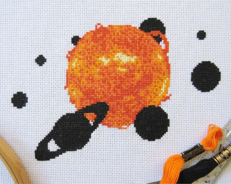 May include: A cross-stitch pattern of a sun with a ring around it, surrounded by smaller black circles. The sun is orange and yellow, and the ring is black. The background is white.