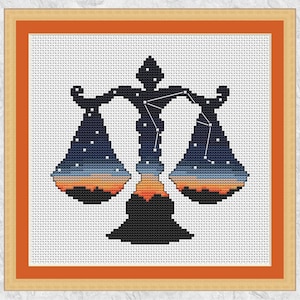 May include: Cross-stitch artwork of the Libra zodiac sign. The scales are black, filled with a gradient of blue, orange, and white, depicting a night sky. The artwork is framed in a wooden frame with an orange border.