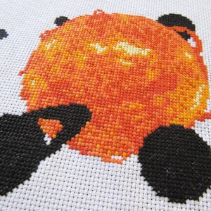 May include: Cross-stitch artwork of a red and orange sun with black details. The sun is surrounded by a white background.