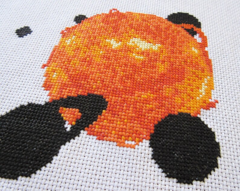 May include: Cross-stitch artwork of a red and orange sun with black details. The sun is surrounded by a white background.